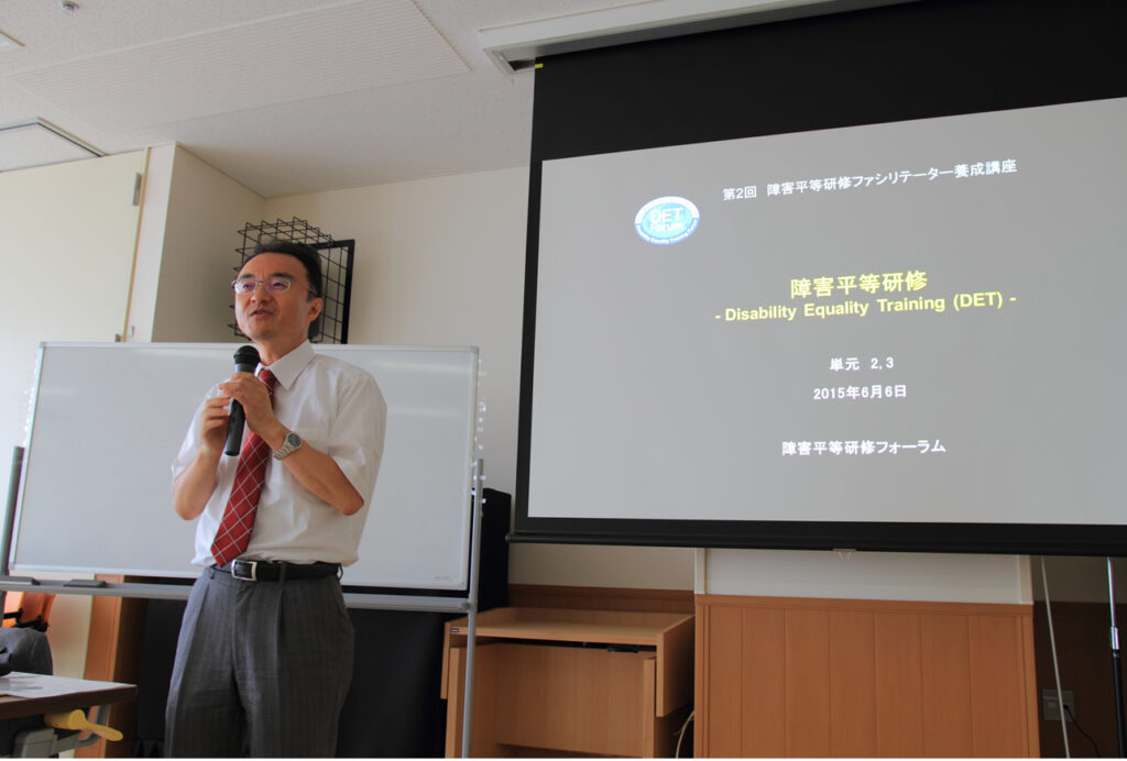 養成講座2回目講師が全体に説明中。講師の右側にはスクリーンありDETのスライドの1ページ目が表示されている
